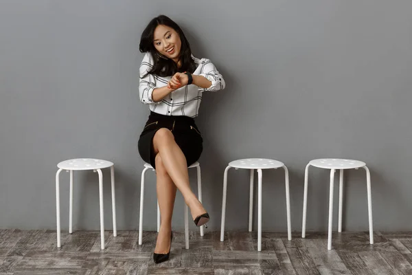 Lächelnde asiatische Geschäftsfrau, die während des Wartens auf ein Vorstellungsgespräch mit dem Smartphone spricht und die Uhrzeit überprüft — Stockfoto