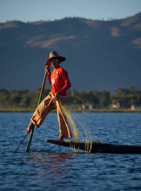 Mandalay, Myanmar. 27 Kasım 2016. Batan güneşin arka planında, Inle Gölü 'nün balıkçısı ağları düzenler, teknenin kenarında durur, aynı zamanda tekneyi kontrol eder, küreği ayağıyla tutar..