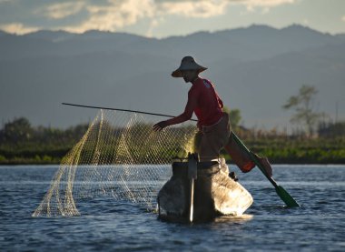 Mandalay, Myanmar. 27 Kasım 2016. Batan güneşin arka planında, Inle Gölü 'nün balıkçısı ağları düzenler, teknenin kenarında durur, aynı zamanda tekneyi kontrol eder, küreği ayağıyla tutar..