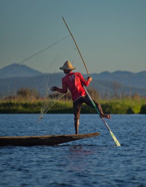 Mandalay, Myanmar. 27 Kasım 2016. Batan güneşin arka planında, Inle Gölü 'nün balıkçısı ağları düzenler, teknenin kenarında durur, aynı zamanda tekneyi kontrol eder, küreği ayağıyla tutar..
