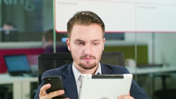 Νεαρός άνδρας χρησιμοποιώντας ένα τηλέφωνο και ένα δισκίο στο γραφείο — Αρχείο Βίντεο