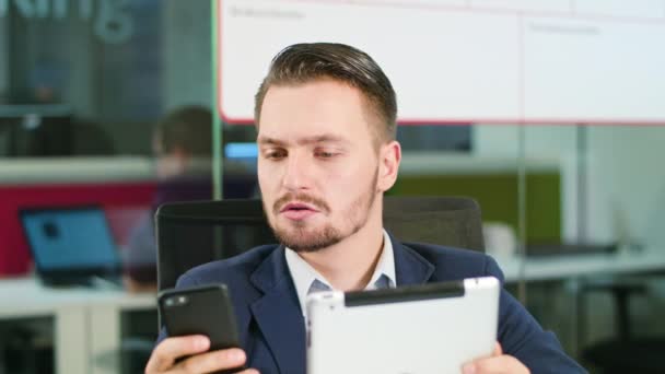 Fiatal férfi használja a telefon és a tabletta az irodában — Stock videók