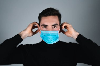 Siyah tişörtlü genç adam izole edilmiş gri arka planda tıbbi maske takıyor.