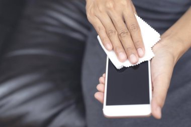 Kanepede oturan bir kadın akıllı telefon ekranını dezenfektanla temizliyor mikrofiber bir bez ve kirli tozdan antiseptik bir sıvı ve diğer dış mikroplar ve bakterilerle. Mobil Bakım Konsepti