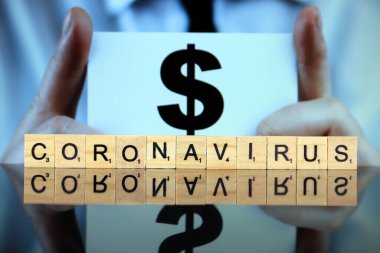 WROCLAW, POLAND - 30 Mart 2020: ahşap harflerden yapılmış CORONAVIRUS sözcüğü, ve arka planda dolar sembolü olan bir kartvizit tutan adam. Coronavirus (COVID-19) küresel hastalık 2020.