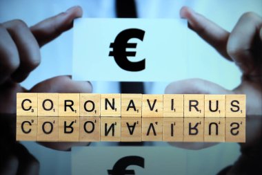 WROCLAW, POLAND - 30 Mart 2020: CORONAVIRUS kelimesi ahşap harflerden yapılmıştır, ve arkasında euro sembolü olan bir kartvizit tutan adam. Coronavirus (COVID-19) küresel hastalık 2020.