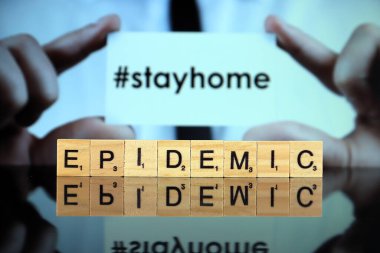 WROCLAW, POLAND - 30 Mart 2020: Ahşap harflerden oluşan EPIDEMIC sözcüğü, ve arka planda STAY HOME (hashtag ile, sosyal medyada popüler) sözleriyle bir kartvizit tutan adam.