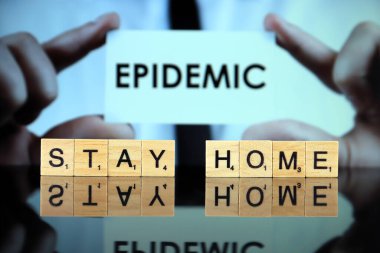 WROCLAW, POLAND - 30 Mart 2020: kelimeler ahşap harflerden yapılmış ev kalır, ve arka planda EPIDEMIC kelimesi olan bir kartvizit tutan adam. Coronavirus (COVID-19) küresel hastalık 2020.