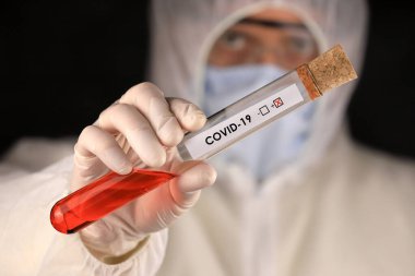 Koruyucu giysili ve maskeli doktor, hızla yayılan Coronavirus 'un pozitif kan testi sonucunu içeren cam bir tüp tutuyor. Küresel koronavirüs hastalığı (COVID-19) 2020.