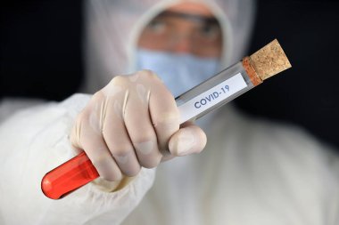 Koruyucu giysili ve maskeli doktor hızla yayılan koronavirüs sırasında üzerinde COVID-19 yazan cam bir tüp tutuyor. Küresel koronavirüs hastalığı (COVID-19) 2020.