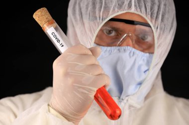 Koruyucu giysili ve maskeli doktor hızla yayılan koronavirüs sırasında üzerinde COVID-19 yazan cam bir tüp tutuyor. Küresel koronavirüs hastalığı (COVID-19) 2020.