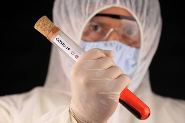 Koruyucu giysili ve maskeli doktor, hızla yayılan Coronavirus 'un pozitif kan testi sonucunu içeren cam bir tüp tutuyor. Küresel koronavirüs hastalığı (COVID-19) 2020.