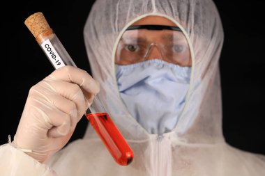 Koruyucu giysili ve maskeli doktor hızla yayılan koronavirüs sırasında üzerinde COVID-19 yazan cam bir tüp tutuyor. Küresel koronavirüs hastalığı (COVID-19) 2020.