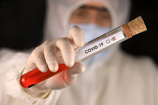 The doctor in a protective suit and mask holding a glass tube with a positive blood test result for the new rapidly spreading Coronavirus. Global coronavirus disease (COVID-19) 2020.