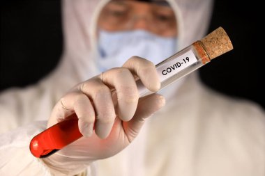 Koruyucu giysili ve maskeli doktor hızla yayılan koronavirüs sırasında üzerinde COVID-19 yazan cam bir tüp tutuyor. Küresel koronavirüs hastalığı (COVID-19) 2020.