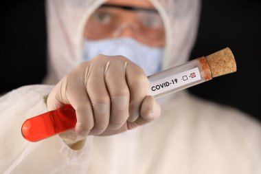 Koruyucu giysili ve maskeli doktor, hızla yayılan Coronavirus 'un pozitif kan testi sonucunu içeren cam bir tüp tutuyor. Küresel koronavirüs hastalığı (COVID-19) 2020.