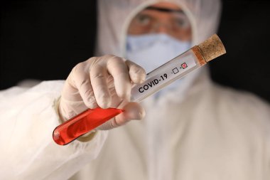 Koruyucu giysili ve maskeli doktor, hızla yayılan Coronavirus 'un pozitif kan testi sonucunu içeren cam bir tüp tutuyor. Küresel koronavirüs hastalığı (COVID-19) 2020.