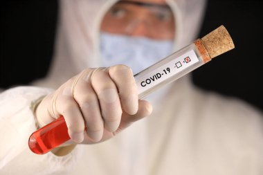 Koruyucu giysili ve maskeli doktor, hızla yayılan Coronavirus 'un pozitif kan testi sonucunu içeren cam bir tüp tutuyor. Küresel koronavirüs hastalığı (COVID-19) 2020.