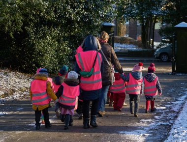 A group of small children, accompanied by adult guardians, walk towards a street in winter attire. A all the children wear high visibility vests and a green first aid kit carried by one of the adults is visible. clipart