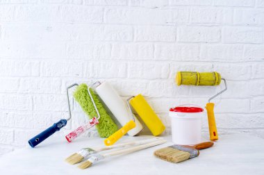 Bir kova boya, sahte tuğlalarla dekore edilmiş beyaz bir duvara karşı beyaz bir masanın üzerinde bir set silindir ve fırçalar..