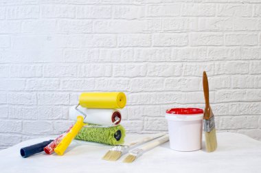 Bir kova boya, sahte tuğlalarla dekore edilmiş beyaz bir duvara karşı beyaz bir masanın üzerinde bir set silindir ve fırçalar..