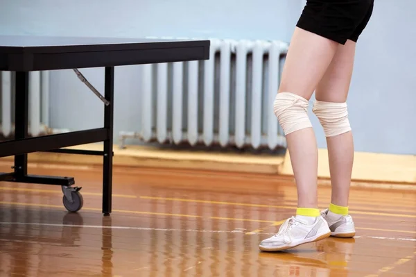 A close-up of a young girl\'s legs is tied with white elastic bandages at the knees next to a tennis table in the gym.