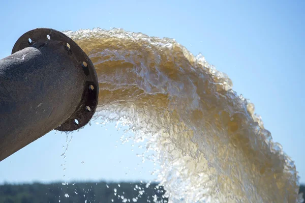 Çamurlu bir su akıntısı borudan mavi gökyüzüne ve uzaktaki yeşil ormana doğru fışkırır..