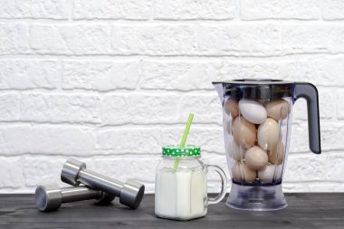 Protein içeceği bardakta, blender kasesinin arka planında tavuk yumurtası ve halterlerle beyaz bir duvara yaslanmış halterler..