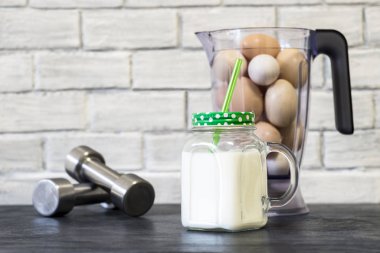 Protein içeceği bardakta, blender kasesinin arka planında tavuk yumurtası ve halterlerle beyaz bir duvara yaslanmış halterler..