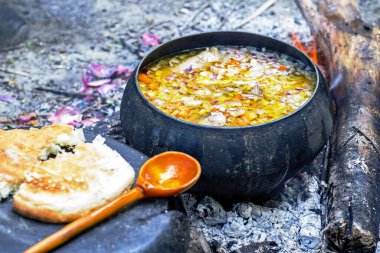 Hazır yemek, şöminenin kömürlerinde antika bir tavada, ön planda tahta kaşık ve beyaz ekmek ile pişiriliyor. Çorbanın keskinliği. Kamp mutfağı konsepti.