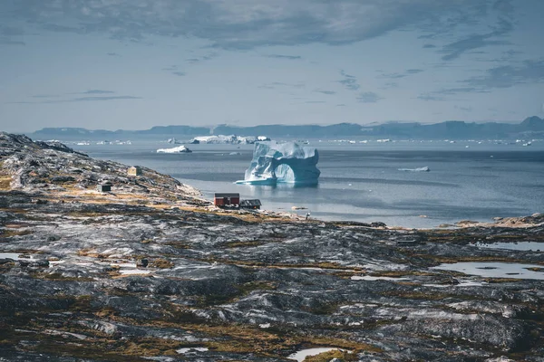 Айсберг з аркою в антарктичних Гренландських водах на тлі гір Арктичного півострова. Маленькі кольорові будинки в сонячний день з блакитним небом і хмарами.. — стокове фото