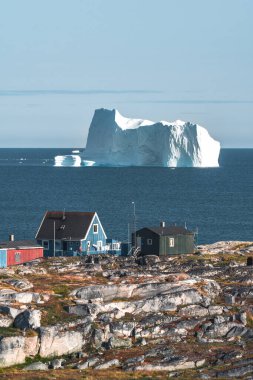 Arka planda buzdağları olan renkli yeşil ahşap evler. Oqaatsut olarak da bilinen Rodebay, Ilulissat 'ın kuzeyinde bir balıkçı kasabasıdır. Disco Bay ve qeqertarsuaq. Mavi gökyüzü ve güneş.