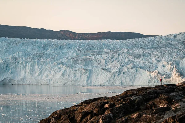 人々は巨大な氷河の氷の壁の前に立って座っている。Eqipグリーンランドのセルミア氷河Eqi氷河は真夜中の太陽の下で氷解する氷河と呼ばれる。旅行や休暇中のハイカー. — ストック写真