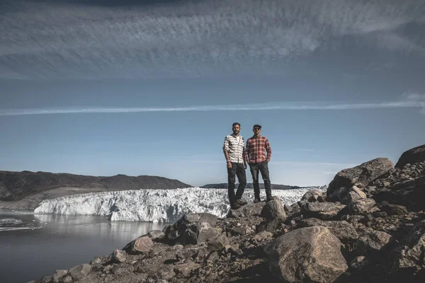 Twee jonge gelukkige mannelijke reiziger die voor de Eqip Sermia Eqi gletsjer in Groenland staat. Uitdagende en vijandige omgeving. Verschillende spleten in de buurt. Blauwe lucht en wolken. Opwarming van de aarde. — Stockfoto