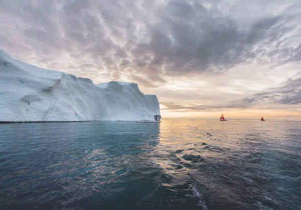 Vacker röd segelbåt i Arktis bredvid ett massivt isberg som visar skalan. Cruising bland flytande isberg i disko Bay Glacier under midnattssol säsongen av Polar Summer Ilulissat, disko Bay — Stockfoto