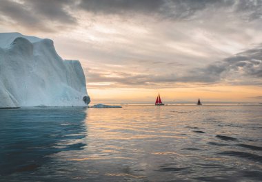 Kuzey kutbunda, ölçeği gösteren devasa bir buzdağının yanında güzel kırmızı yelkenli. Polar yaz Ilulissat, Disko Körfezi gece yarısı güneş sezonu sırasında Disko Körfezi buzul yüzen buzdağları arasında seyir
