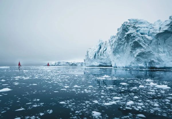 북극의 아름다운 붉은 범선은 규모를 보여주는 거대한 빙산 옆에 있습니다. 극지 여름 일룰리사트, 디스코 베이의 한밤중 태양 절기 동안 디스코 베이 빙하의 떠다니는 빙산 사이를 순항 — 스톡 사진