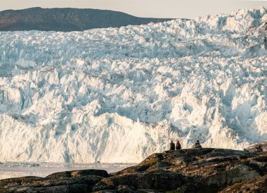 İnsanlar buzdan dev bir duvarın önünde oturuyorlar. Grönland 'daki Eqip Sermia Buzulu Eqi buzuluna gece yarısı güneşi altında buzul yavrusu denir. Gezi ve tatil sırasında yürüyüşçüler.