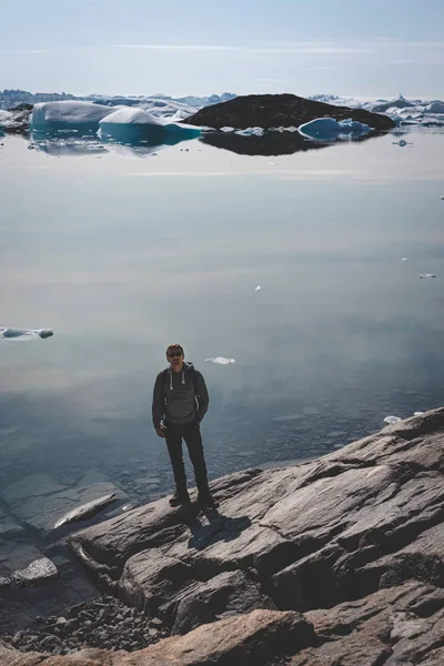 Pohled na Icefjord v Ilulissat. Mladý muž cestovatel turista stojí na promenádě. Ledovce z ledovce Kangia v Grónsku plavou s modrou oblohou a mraky. Symbol globálního oteplování. — Stock fotografie