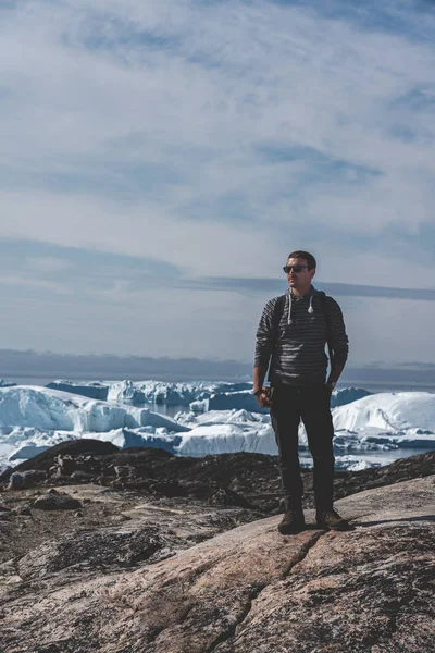 Kilátás Icefjord felé Ilulissatban. Fiatalember utazó turista áll a sétányon. Jéghegyek a grönlandi Kangia gleccserből, kék égbolttal és felhőkkel úszva. A globális felmelegedés szimbóluma. — Stock Fotó