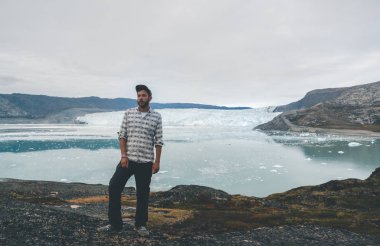 Eqip Sermia buzulunun önünde duran mutlu ve gülümseyen genç adam Eqi Buzulu. Arka planda kocaman buz duvarı olan bir turist. Küresel ısınma ve profesyonel rehberlik kavramı. Bulutlu
