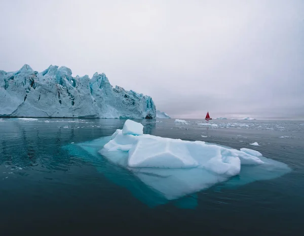 Vacker röd segelbåt i Arktis bredvid ett massivt isberg som visar skalan. Cruising bland flytande isberg i disko Bay Glacier under midnattssol säsongen av Polar Summer Ilulissat, disko Bay — Stockfoto