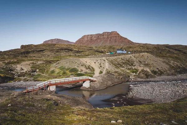 Sentiero escursionistico sull'isola artica di Disko in Groenlandia. Letto fluviale con ponte e case colorate sullo sfondo. Montagne da tavola in una giornata di sole in estate . — Foto Stock