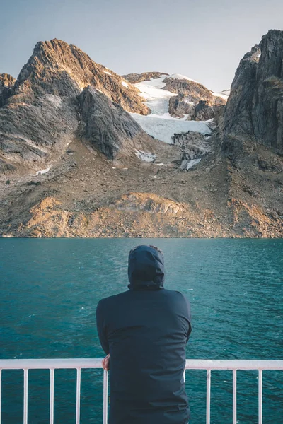 Joven congelado de pie en la cubierta de un barco en el Ártico con montañas cubiertas de nieve en el fondo. A bordo del buque costero Sarfaq Ittuk en Groenlandia que viaja de Ilulissat a Nuuk . —  Fotos de Stock