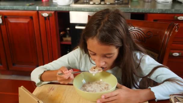 8 〜 10 歳の女の子は台所のテーブルに牛乳とコーンフレークを食べるし、彼女のスマート フォンに見えます。4 k、スローモーション — ストック動画