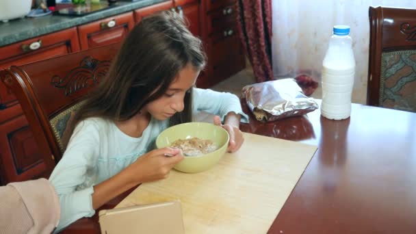 Une fille de 8 à 10 ans mange des céréales pour petit déjeuner avec du lait à une table dans la cuisine et regarde dans son smartphone. 4k, au ralenti — Video