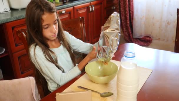 Une fille de 8 à 10 ans mange des céréales pour petit déjeuner avec du lait à une table dans la cuisine et regarde dans son smartphone. 4k, au ralenti — Video