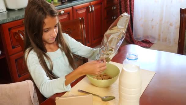 Una niña de 8-10 años come cereales de desayuno con leche en una mesa en la cocina y mira en su teléfono inteligente. 4k, cámara lenta — Vídeo de stock