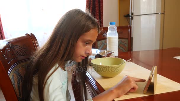 Une fille de 8 à 10 ans mange des céréales pour petit déjeuner avec du lait à une table dans la cuisine et regarde dans son smartphone. 4k, au ralenti — Video