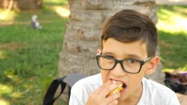 Çekici çocuk çim Park'ta bir ağacın altında oturup taze hamburger yeme. 4k, ağır çekim